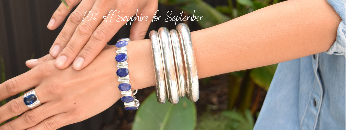A person's wrist and hand wearing a silver bangle bracelet with deep blue sapphire stones, a matching silver ring, and a stack of thick silver bangles
